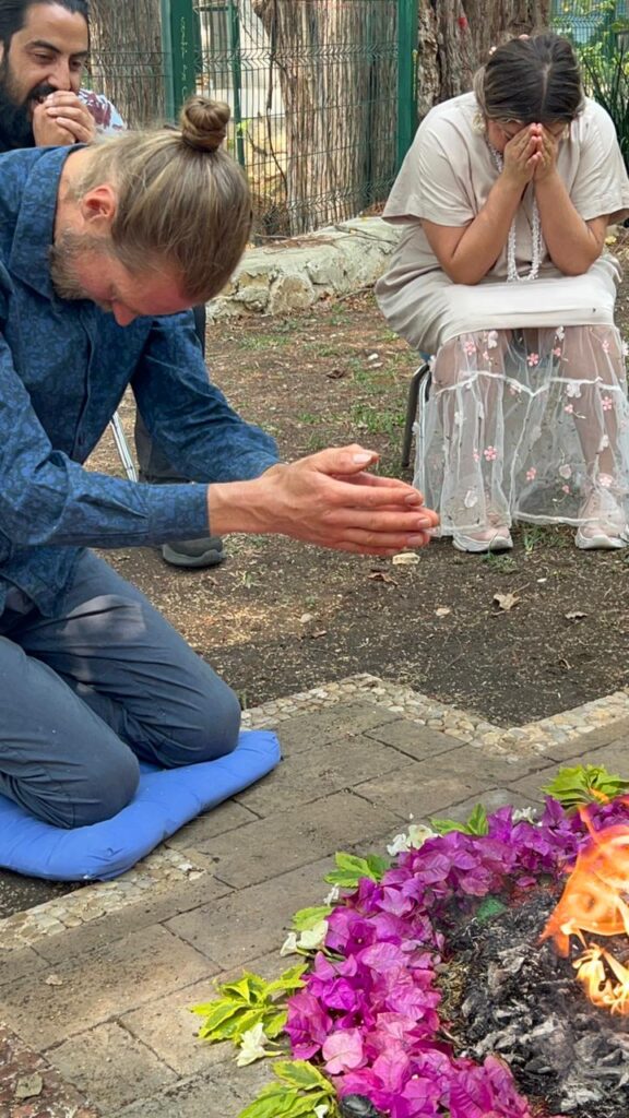 Joakim kneeling in prayer at a Maya fire ceremony, hands extended toward ceremonial fire surrounded by flowers
